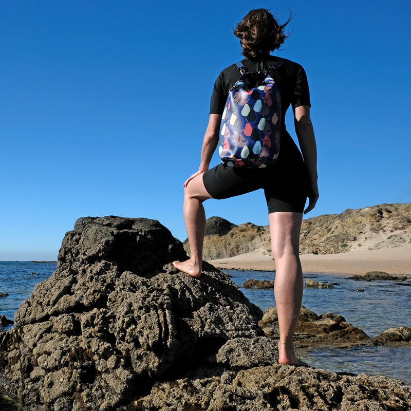 Frau steht mit dem Drybag Peras blau auf dem Rücken auf einem Felsen am Meer