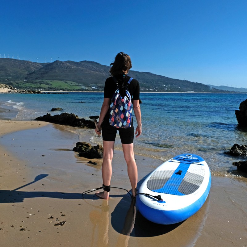 Frau trägt den Drybag Peras blau am Strand neben einem SUP-Board
